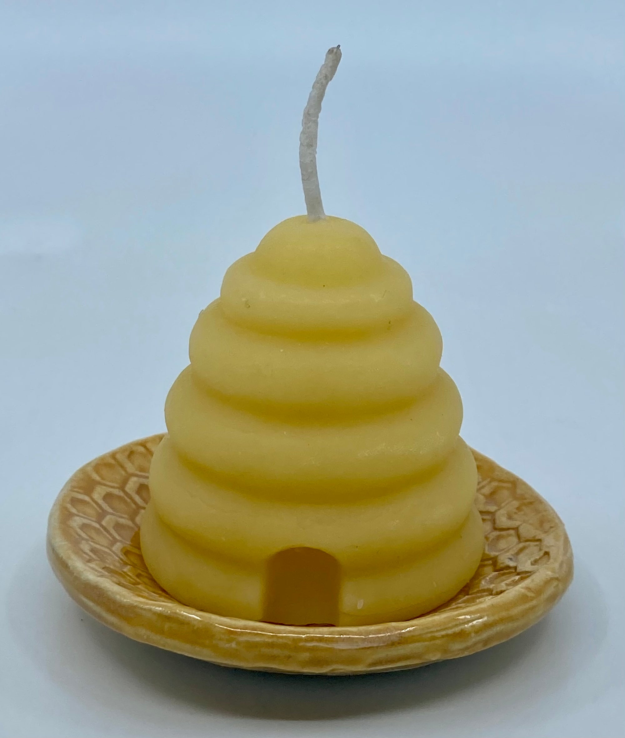 Beeswax candle in the shape of an old fashioned bee hive, which is also known as a skep.  The candle is sitting on a small ceramic dish with a honeycomb pattern on it.